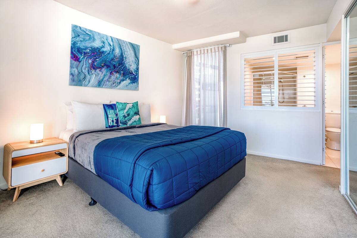 A bedroom with a bed, a nightstand, a wall-mounted artwork, and a window with blinds.