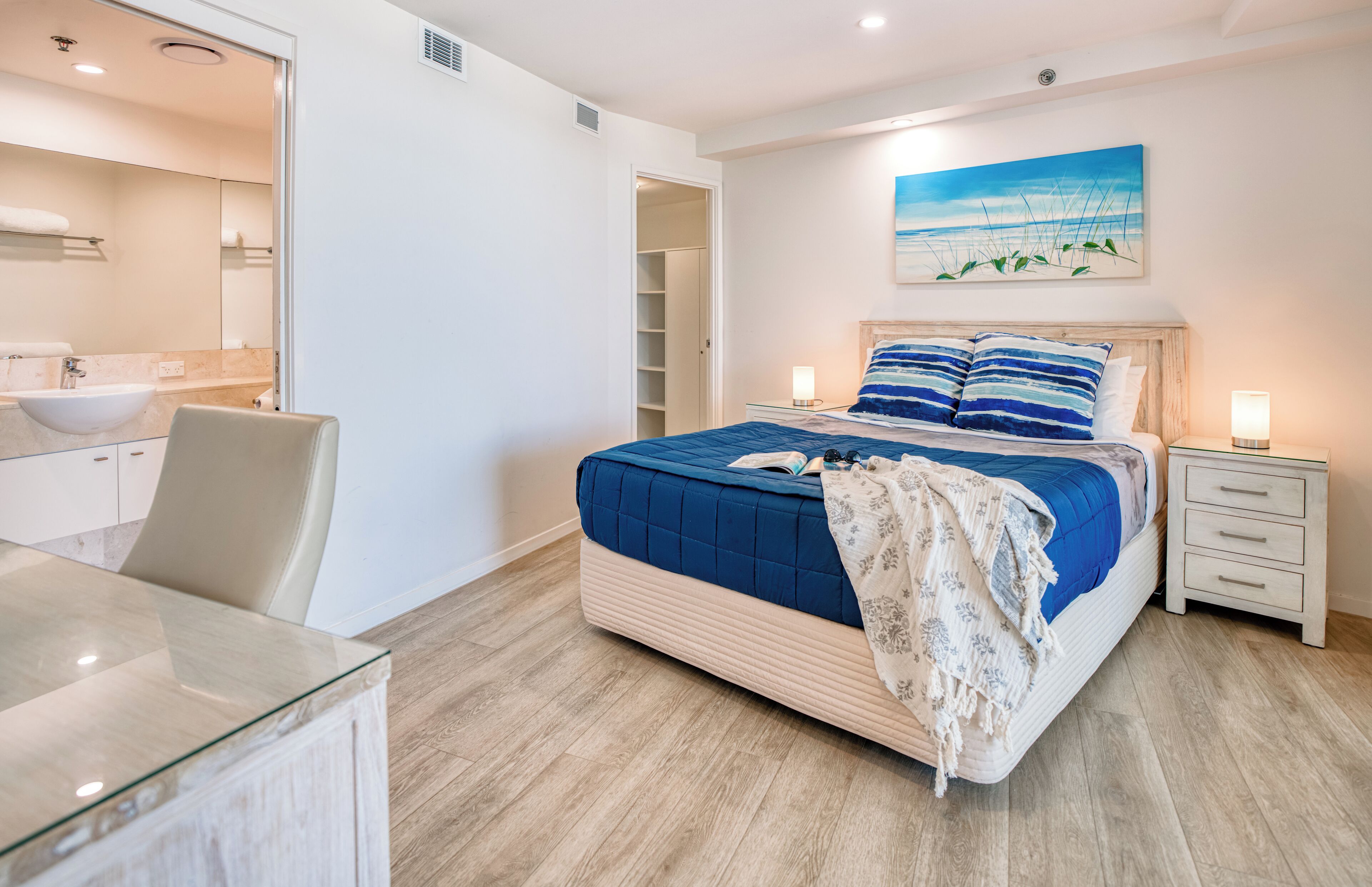 Une chambre à coucher moderne avec un grand lit, des tables de chevet, une table en verre, une salle de bain avec un lavabo et un miroir, et un tableau accroché au mur.