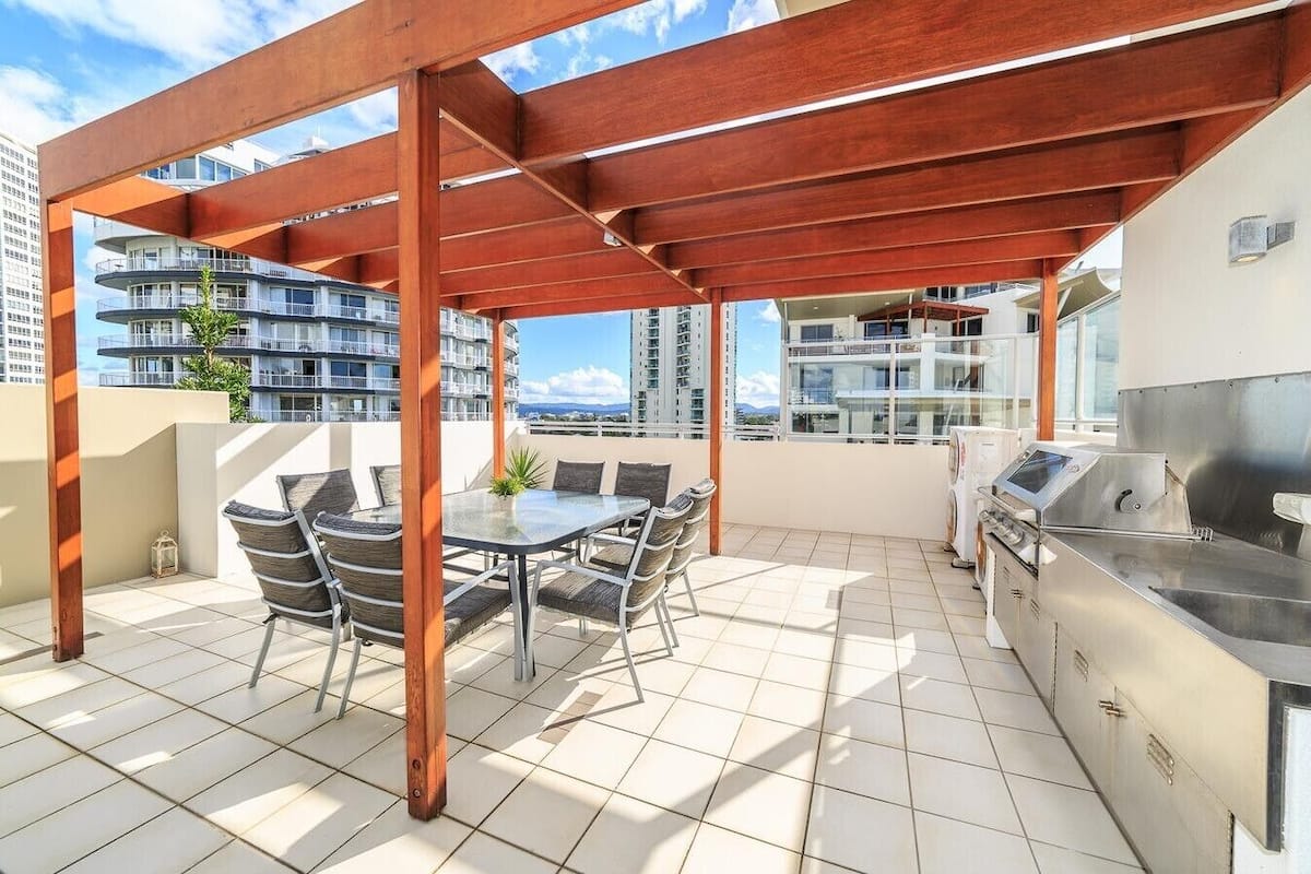 A rooftop patio with a wooden pergola, a dining table with chairs, and a stainless steel grill.