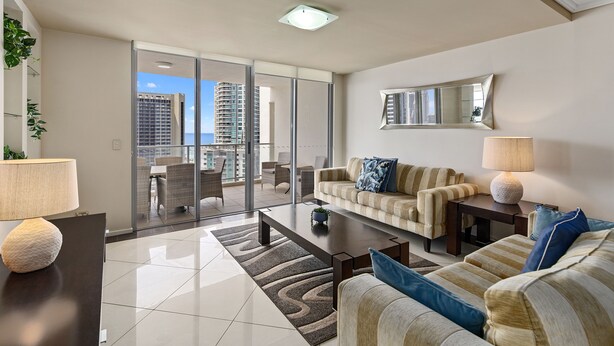 A modern living room with a sofa, coffee table, and lamps. There is a large window with a view of tall buildings. - Trilogy Surfers Paradise (Surfers Paradise)