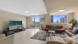 A modern living room with a large flat-screen TV, leather sofas, a coffee table, and a view of the city through large windows. - Trilogy Surfers Paradise (Surfers Paradise)