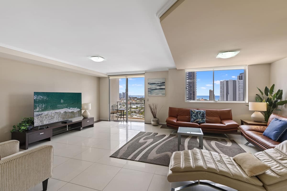 A modern living room with a large flat-screen TV, leather sofas, a coffee table, and a view of the city through large windows.