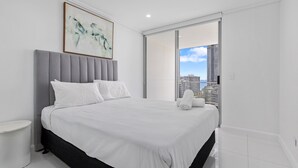 A modern bedroom with a large bed, a grey upholstered headboard, a white bedside table, and a view of a cityscape through a sliding glass door. - Trilogy Surfers Paradise (Surfers Paradise)
