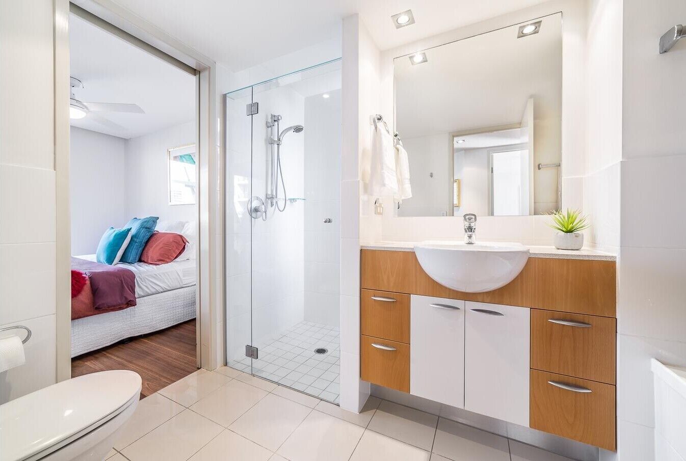 A modern bathroom with a glass shower enclosure, a white sink with a wooden vanity, and a visible bed with colorful pillows in the adjacent room.