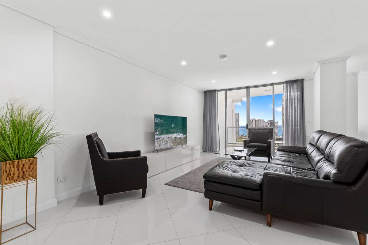 A modern living room with a black leather sofa, a black armchair, and a glass coffee table. There is a large window with a city view, and a painting of a beach scene on the wall.