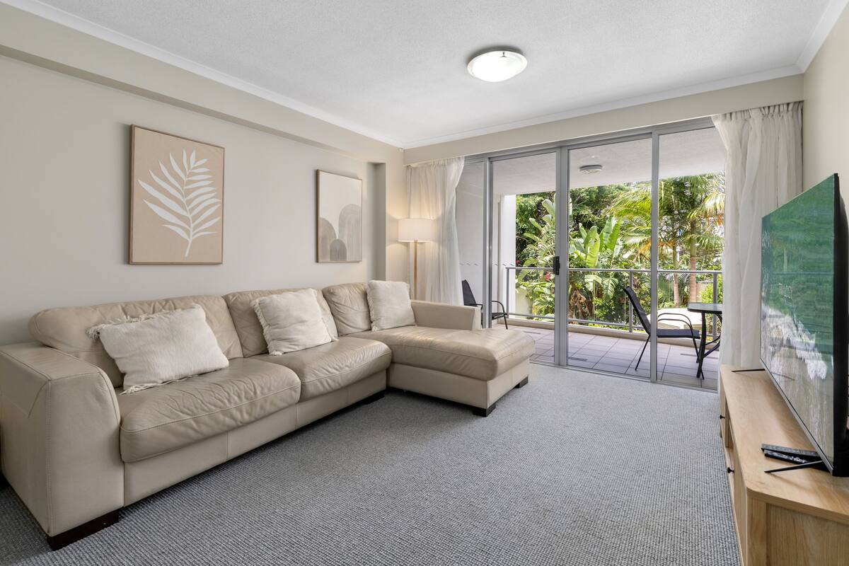 A modern living room with a beige sectional sofa, a flat-screen TV on a wooden stand, and a sliding glass door leading to an outdoor area with greenery.