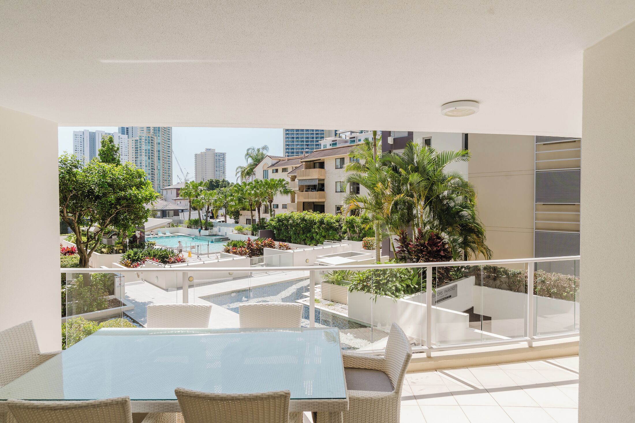 Uma varanda com mesa de vidro e cadeiras de vime, com vista para uma piscina e vegetação, com edifícios altos ao fundo.