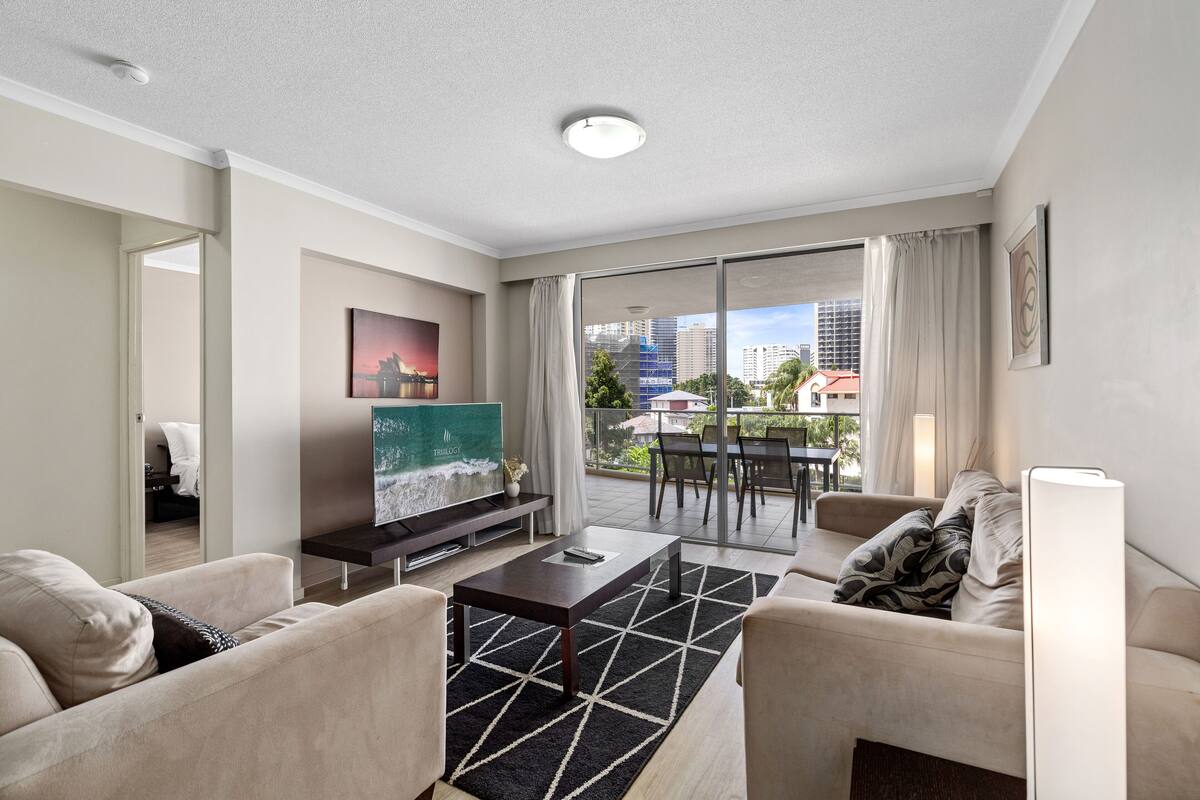 A modern living room with a sofa, coffee table, and a flat-screen TV. There is a balcony with a view of the city.