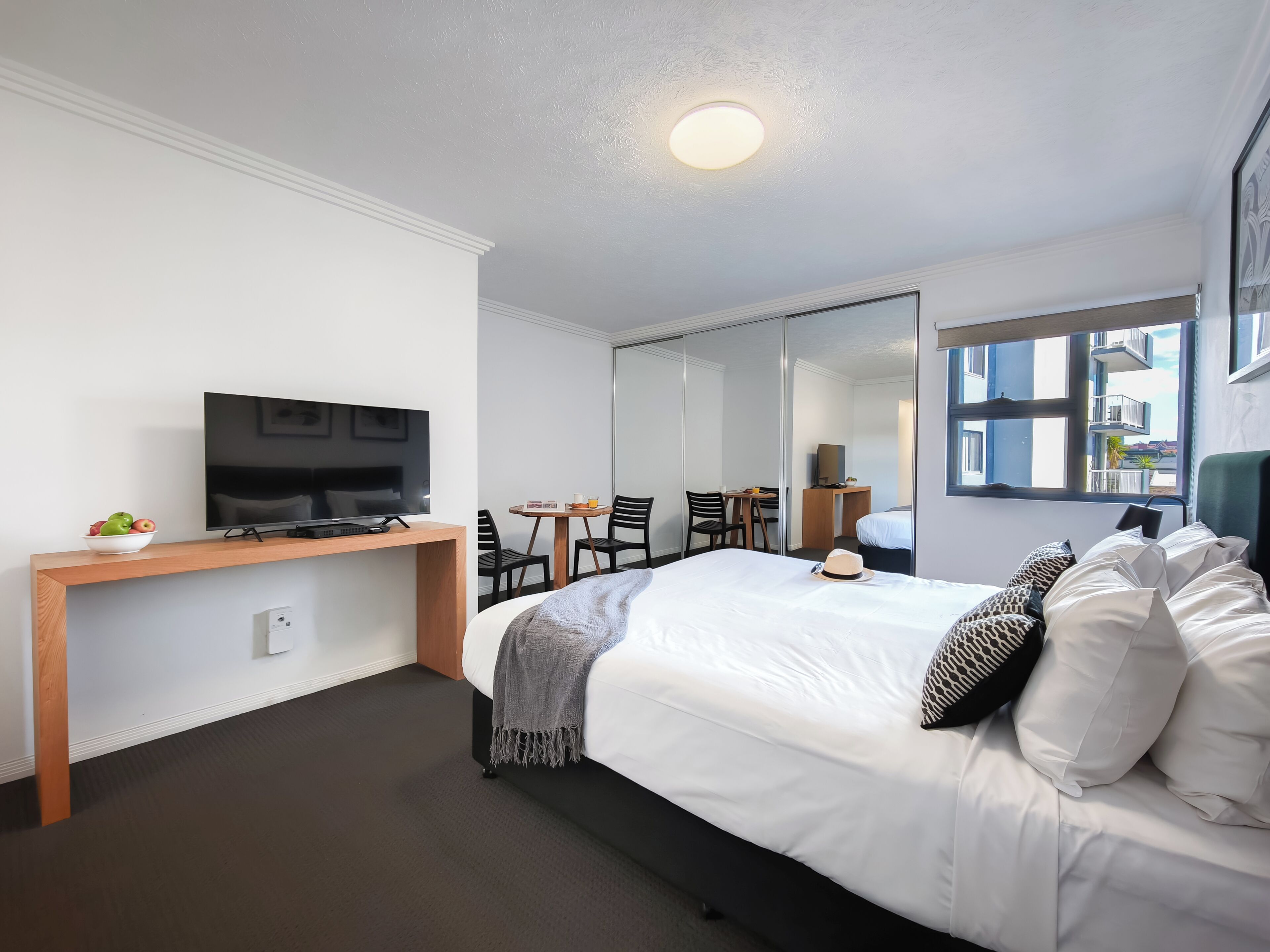 Habitación de hotel con cama, escritorio con televisor, una mesita con sillas, espejo y ventana con vistas.