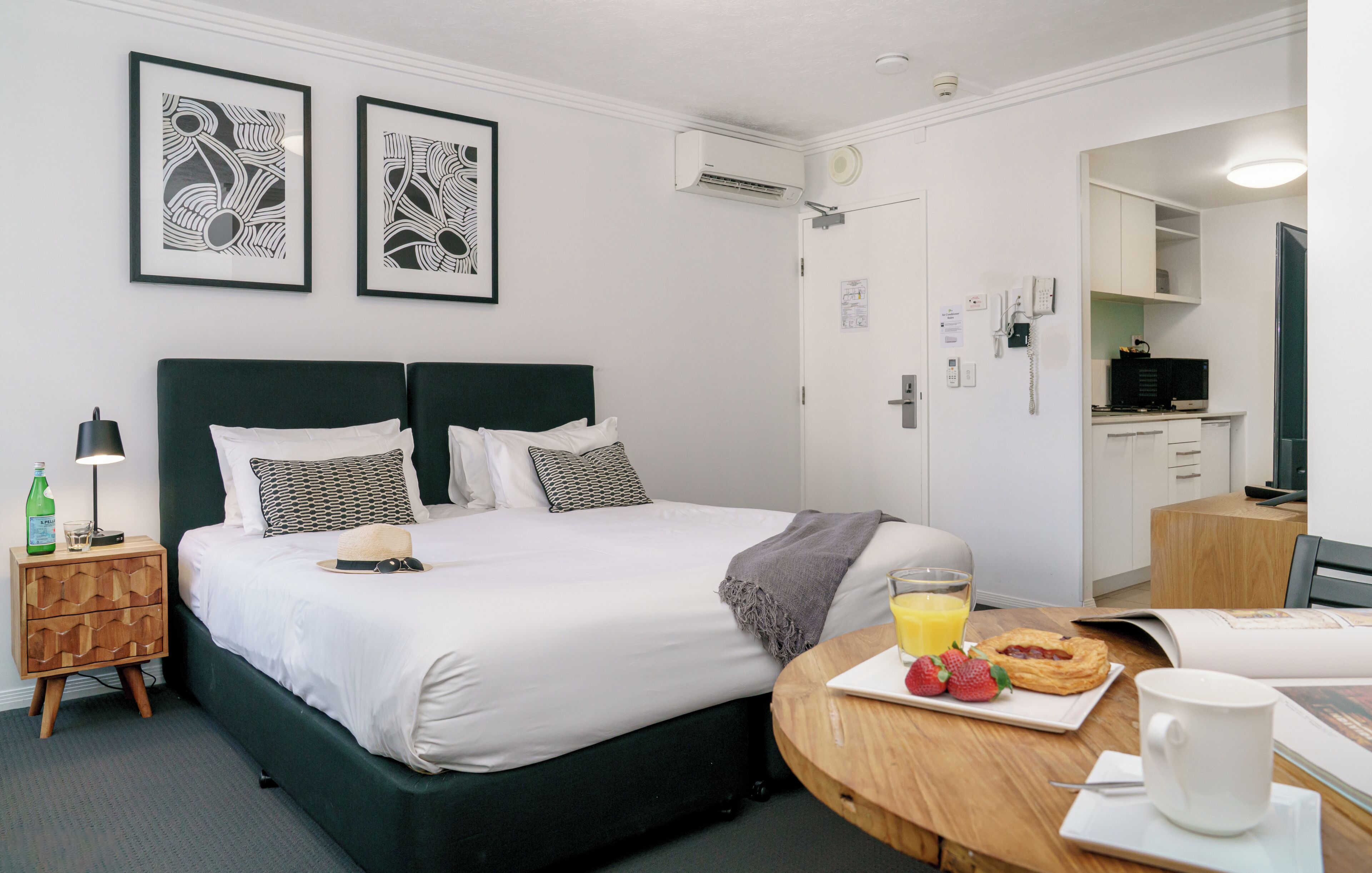 A hotel room with a bed, bedside table, lamp, artwork, kitchenette, and breakfast items.