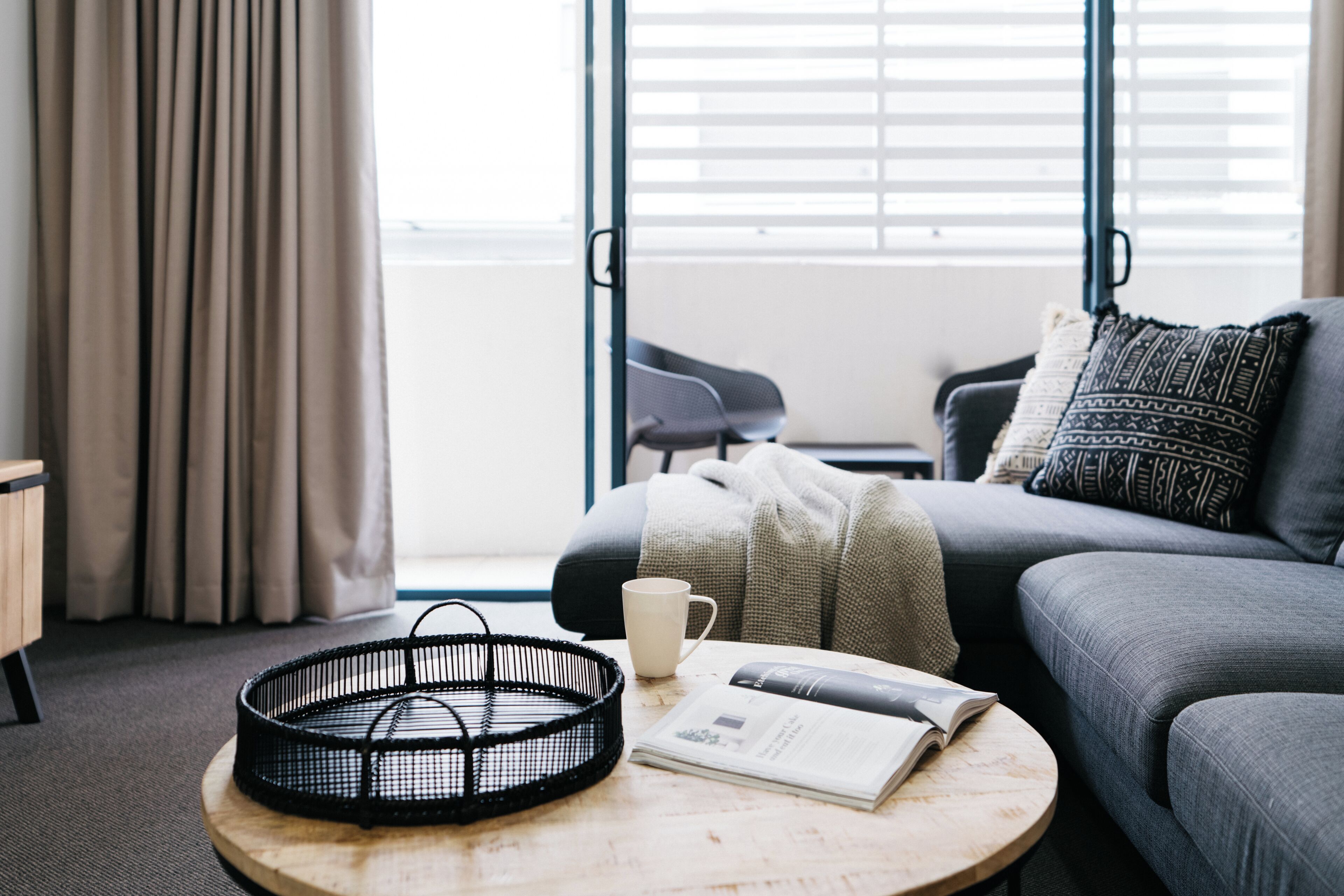 A modern living room with a round wooden coffee table, a black woven basket, a white mug, and a magazine.
