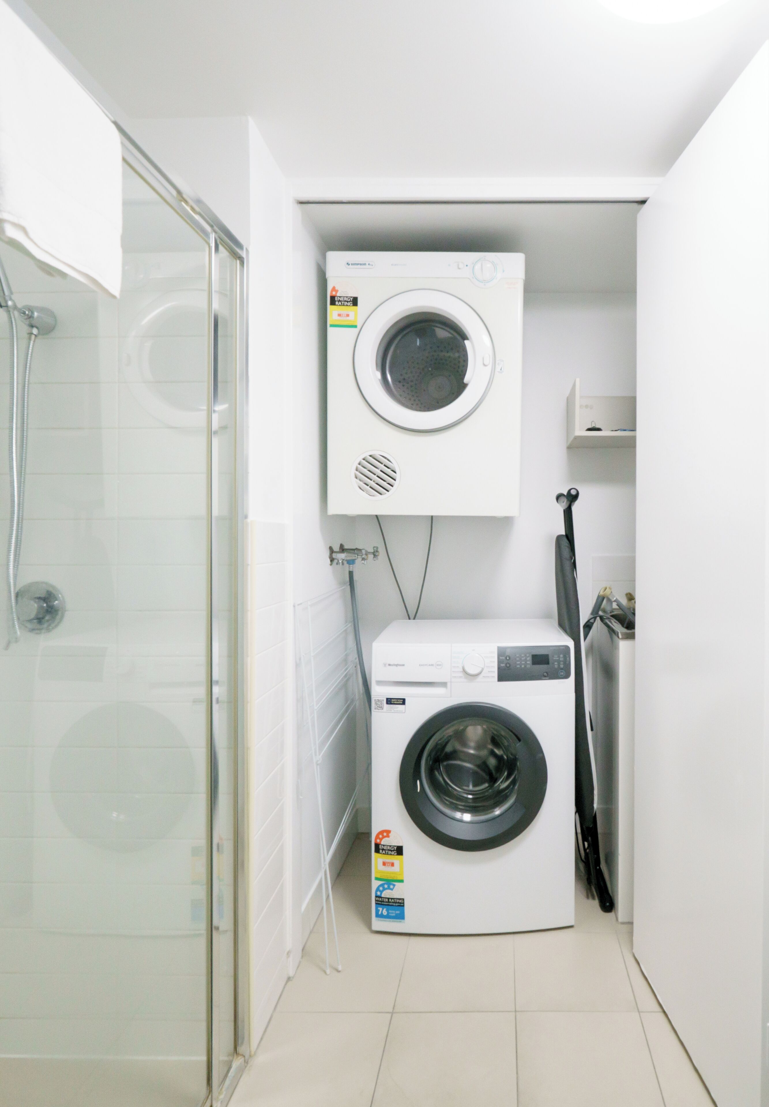 A compact laundry room with a front-loading washing machine and a dryer, a glass shower enclosure, and a small shelf above the dryer.