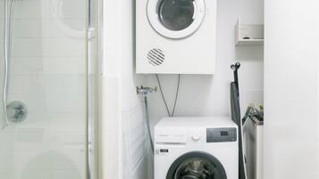 A compact laundry room with a front-loading washing machine and a dryer, a glass shower enclosure, and a small shelf above the dryer.