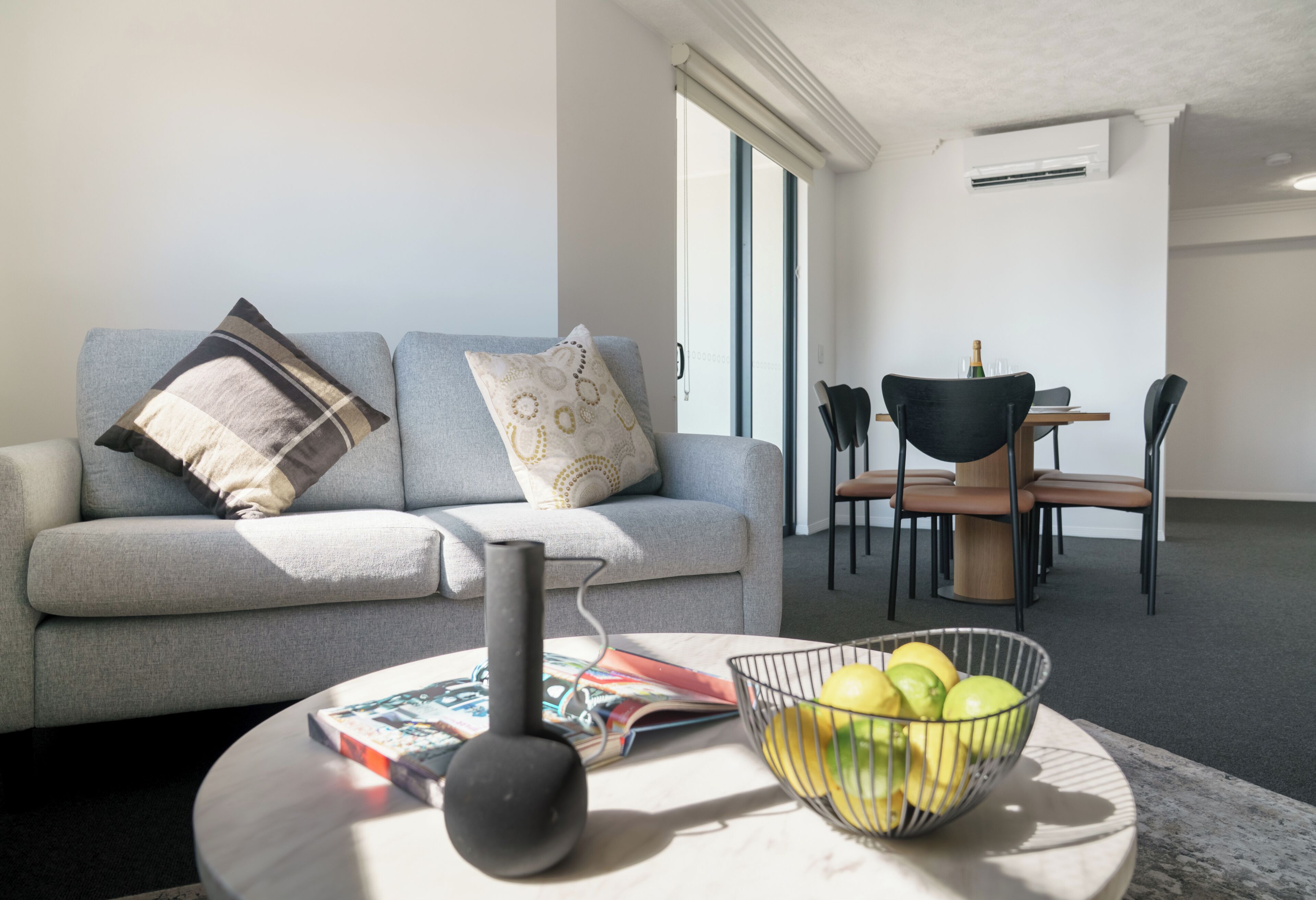 A modern living room with a grey sofa, a coffee table with a magazine and a speaker, and a dining area with a table and chairs.