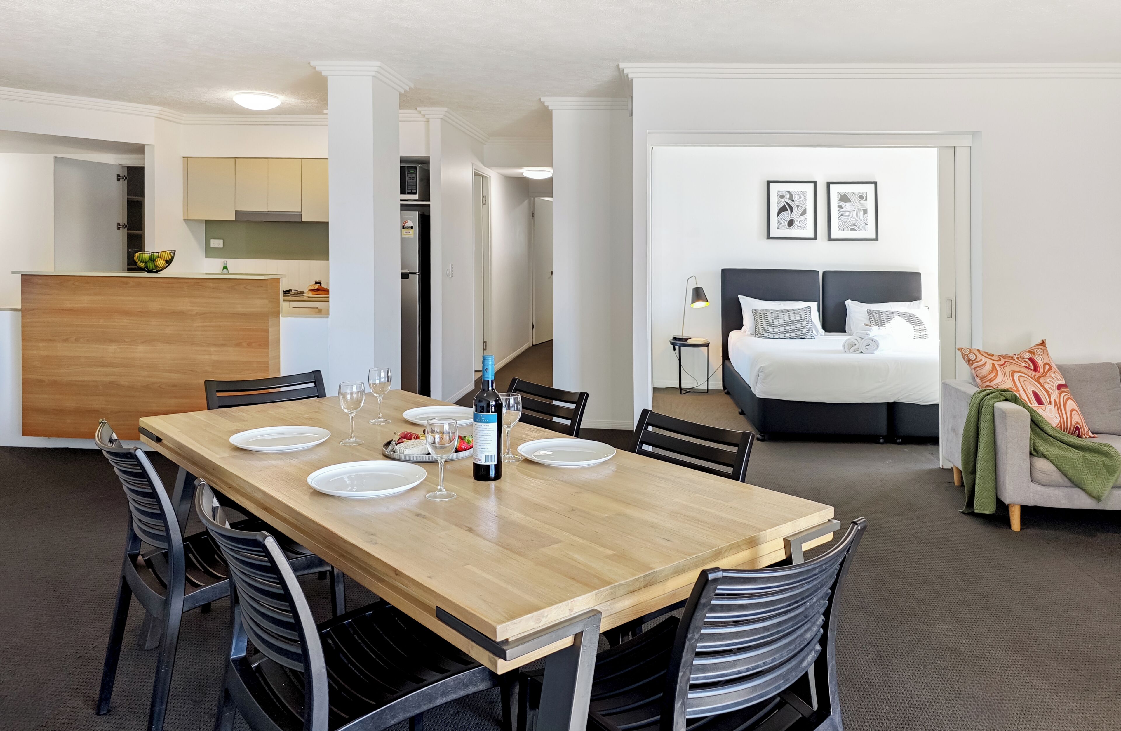 A modern hotel room with a dining area, kitchenette, and a bed with a headboard.
