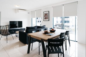 A modern living room with a dining area, a flat-screen TV, and a large window with blinds.