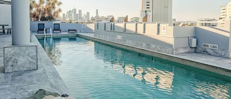 A rooftop pool with clear blue water, a lounge area with chairs, and a city skyline in the background.