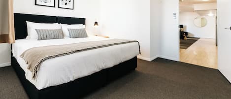 Una habitación de hotel con una cama grande, cabecera, dos mesitas de noche con lámparas, piso alfombrado y una puerta que da a un pasillo.
