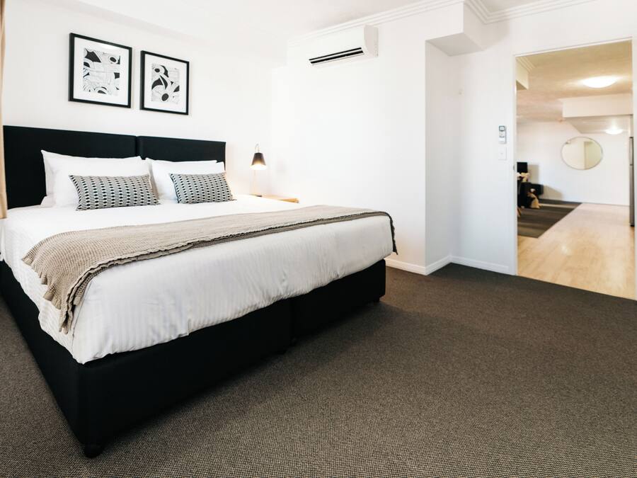 Una habitación de hotel con una cama grande, cabecera, dos mesitas de noche con lámparas, piso alfombrado y una puerta que da a un pasillo.