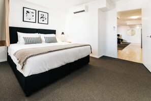 A hotel room with a large bed, a headboard, two bedside tables with lamps, a carpeted floor, and a door leading to a hallway.