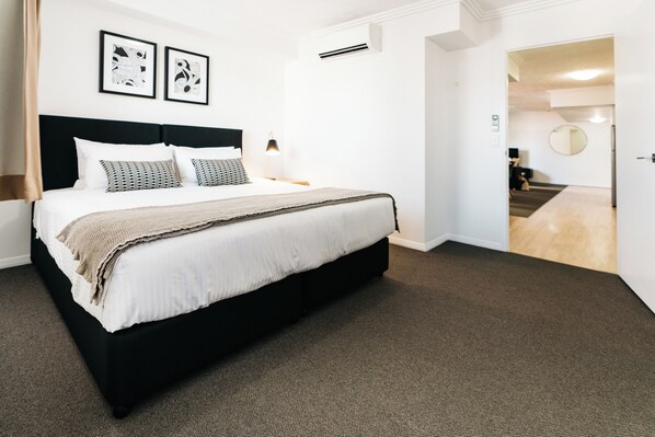 A hotel room with a large bed, a headboard, two bedside tables with lamps, a carpeted floor, and a door leading to a hallway.