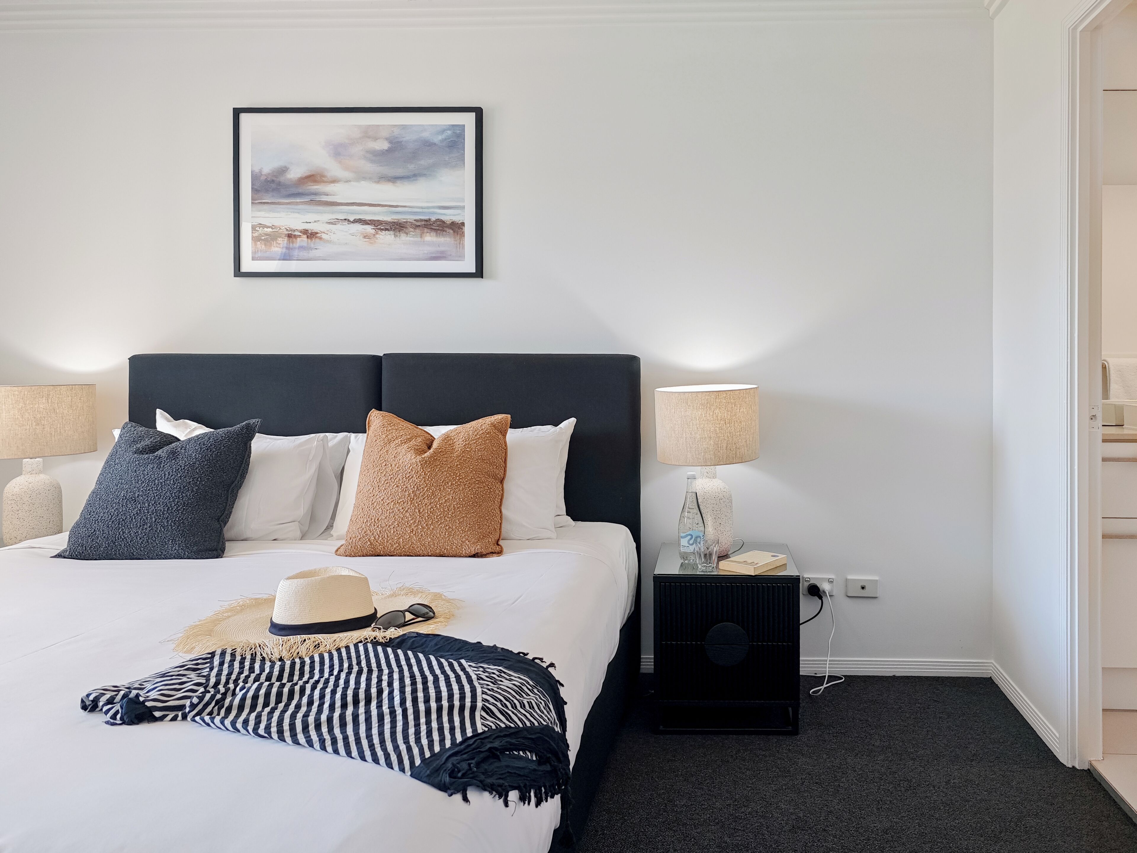 A neatly made bed with a headboard, two bedside lamps, a framed picture on the wall, and a small bedside table with a lamp and a bottle.