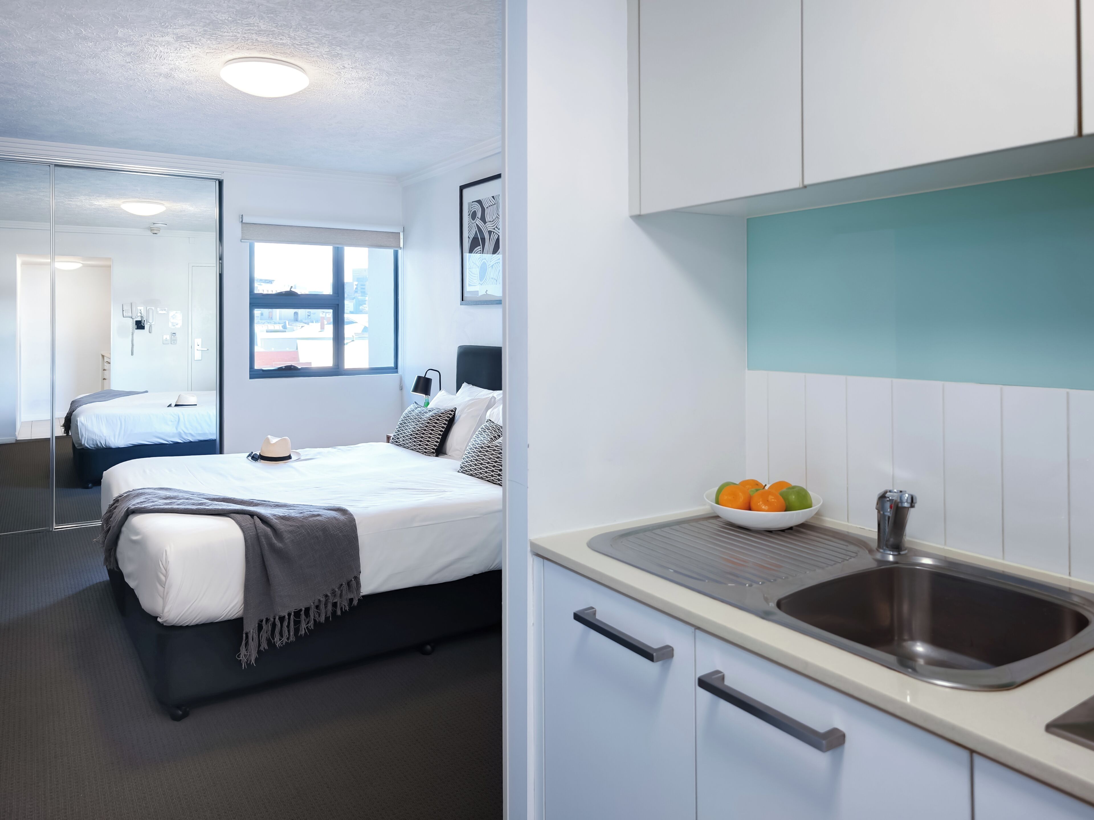 Una habitación de hotel moderna con una cama, una mesita de noche, un espejo, una ventana y una kitchenette con un lavabo y un cuenco de fruta.