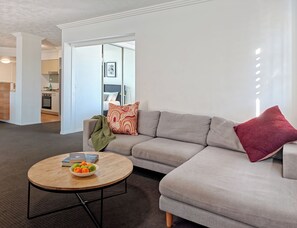 A modern living room with a grey sectional sofa, a round wooden coffee table with fruits, and a view into the kitchen. - Gabba Central Apartments (Woolloongabba)