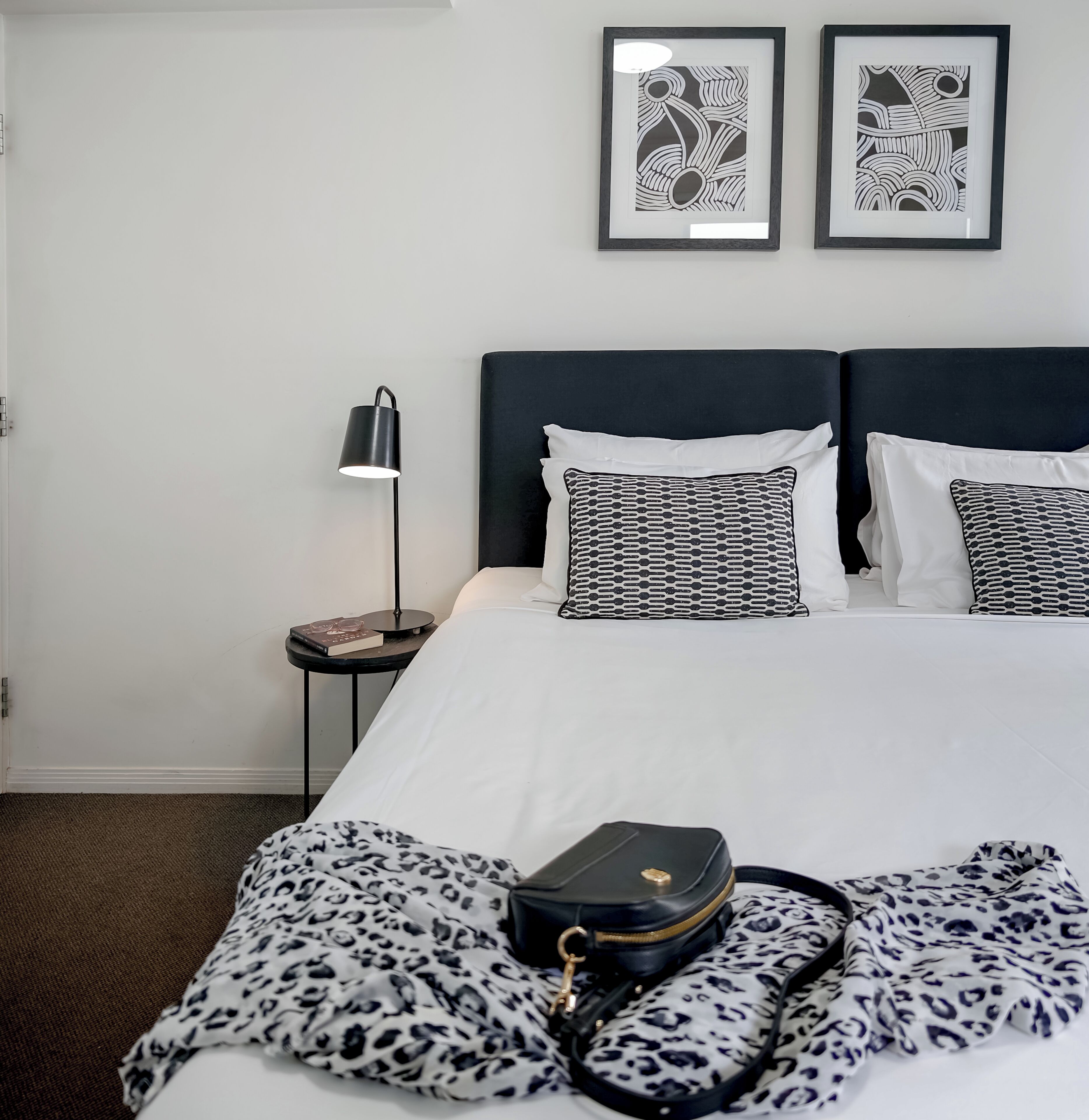 A hotel room with a bed, two pillows, a nightstand with a lamp, and a framed abstract artwork on the wall.