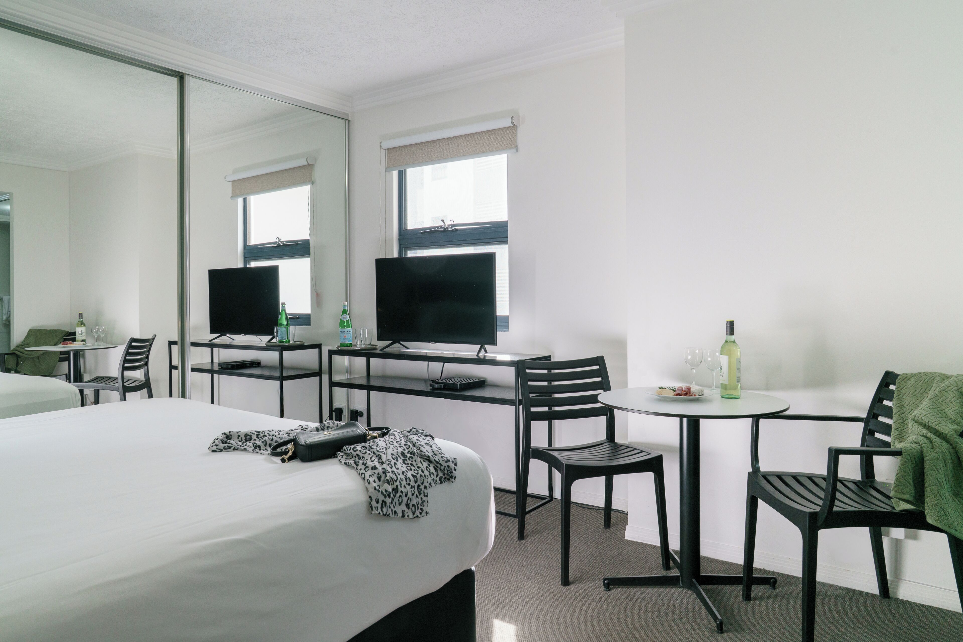 A hotel room with a bed, a desk with a television, a chair, a small table with a bottle and glasses, and a mirror.