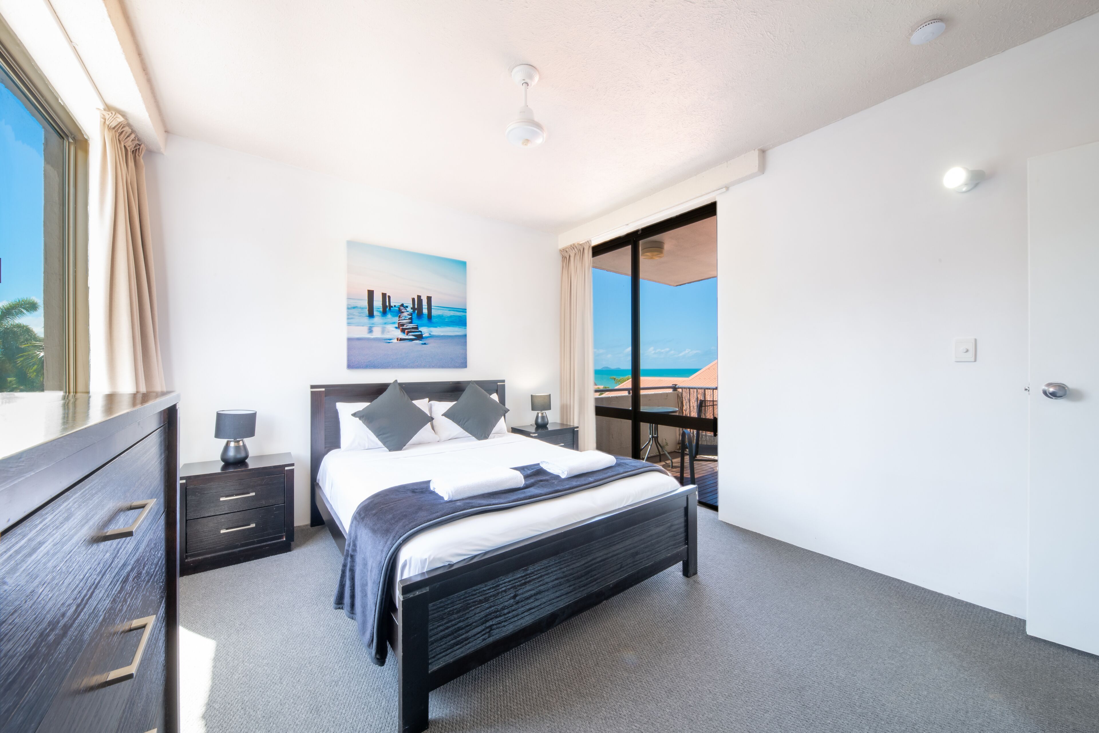 A modern bedroom with a large bed, a nightstand, a dresser, and a view of the ocean through a sliding door.