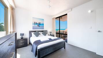 A modern bedroom with a large bed, a nightstand, a dresser, and a view of the ocean through a sliding door.