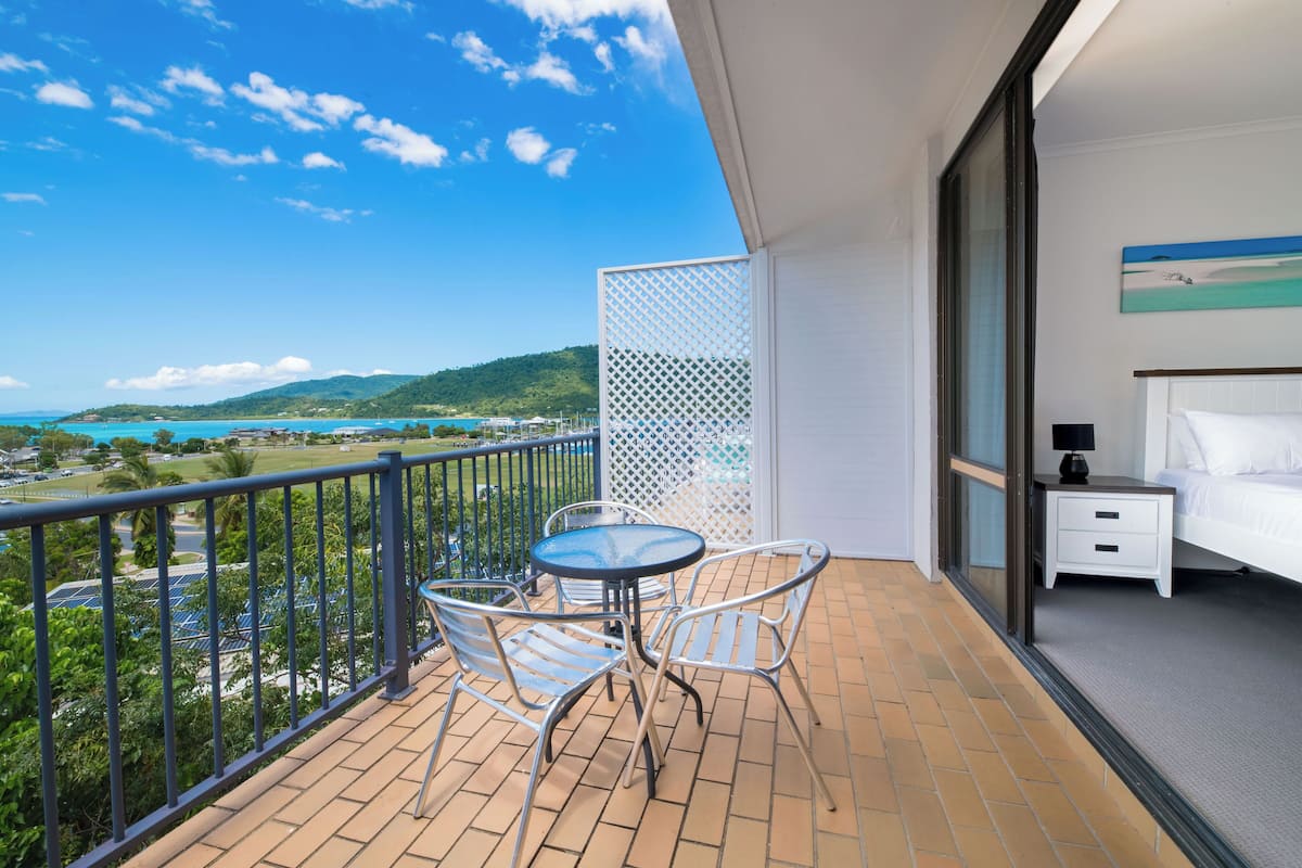 A balcony with a table and chairs, a bed, and a view of a coastal landscape.