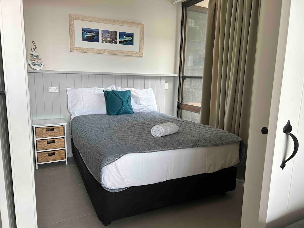 A neatly made bed with a grey and white duvet, a teal pillow, and a white pillow. There is a small bedside table with drawers and a framed picture above the bed.