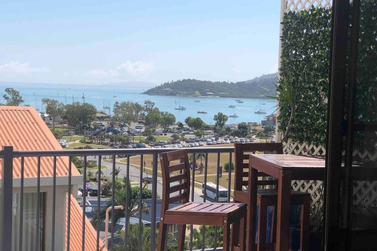 A balcony with wooden chairs and a table, overlooking a marina with boats and a distant island.
