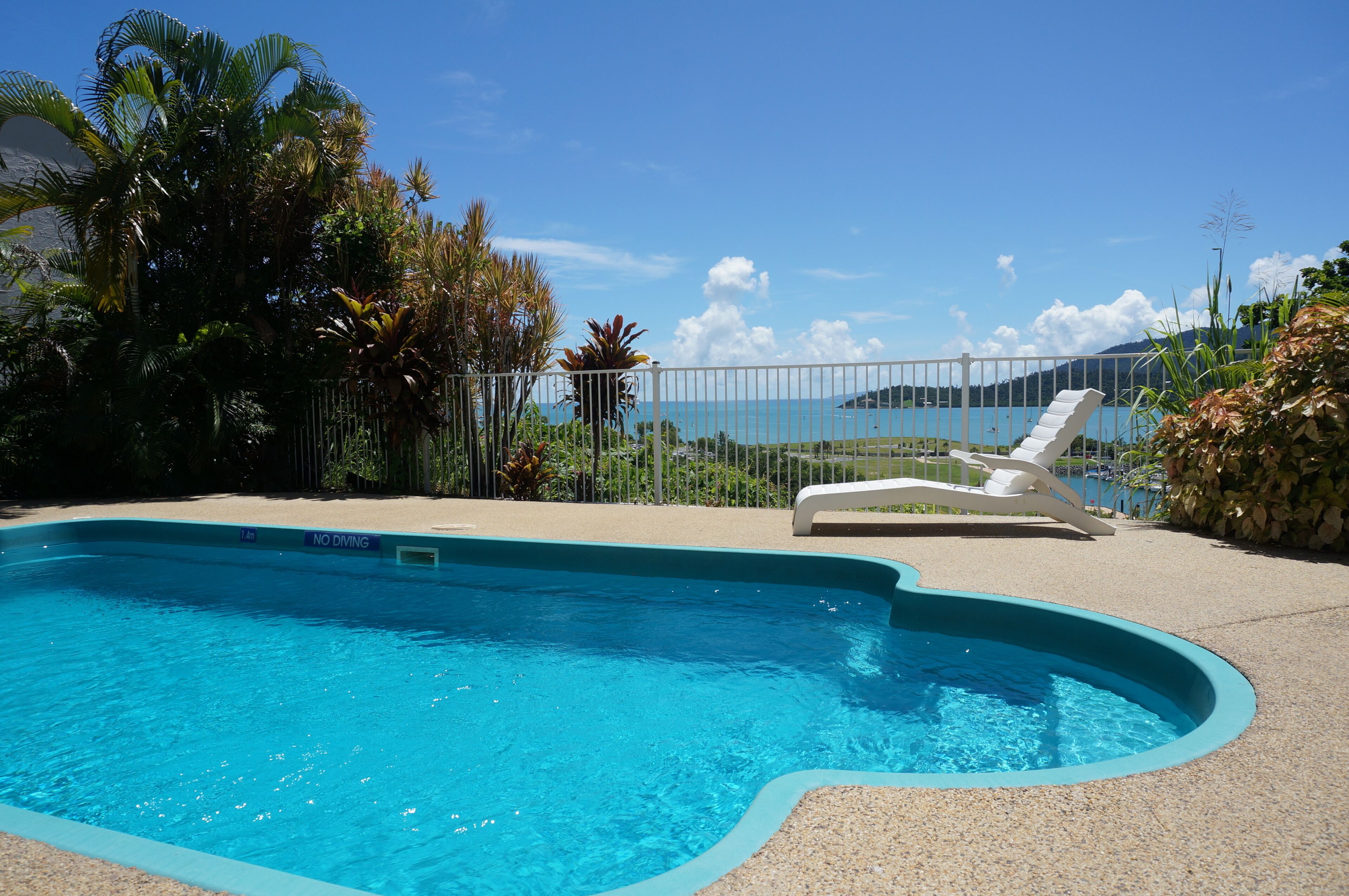 A swimming pool with a 'NO DIVING' sign, surrounded by a fence, with a view of the sea and mountains in the background.