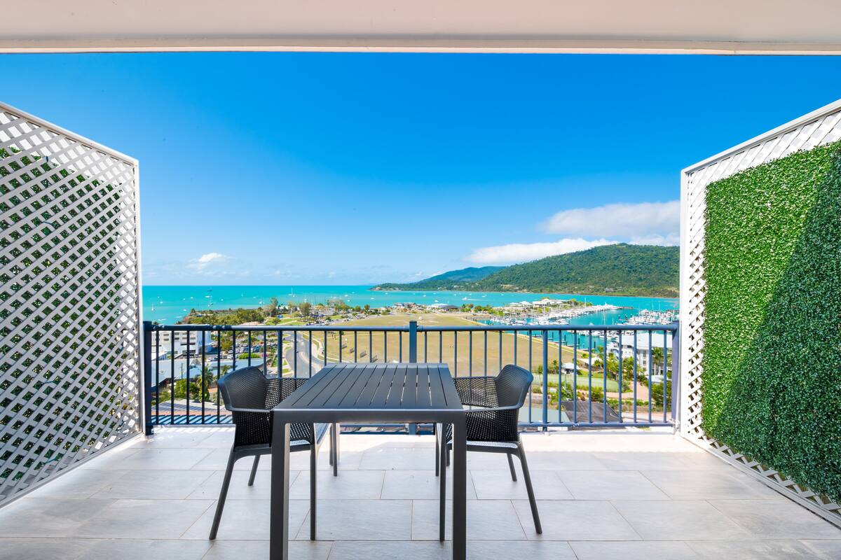 A balcony with a table and chairs, overlooking a beach and marina with clear blue waters and a hilly landscape in the distance.