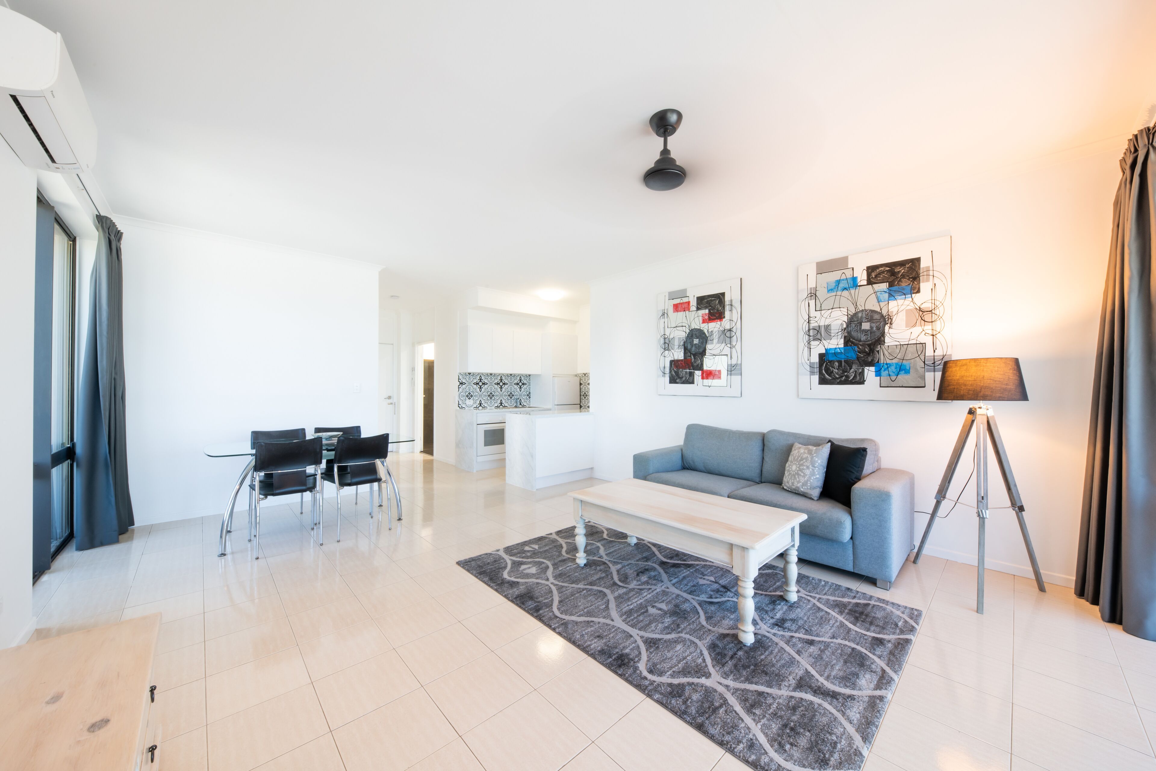 Een moderne woonkamer met een bank, salontafel en een eethoek. Achtergrond is een keuken en er hangt een plafondventilator.