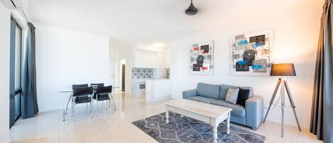 A modern living room with a sofa, coffee table, and dining area. There is a kitchen in the background and a ceiling fan.