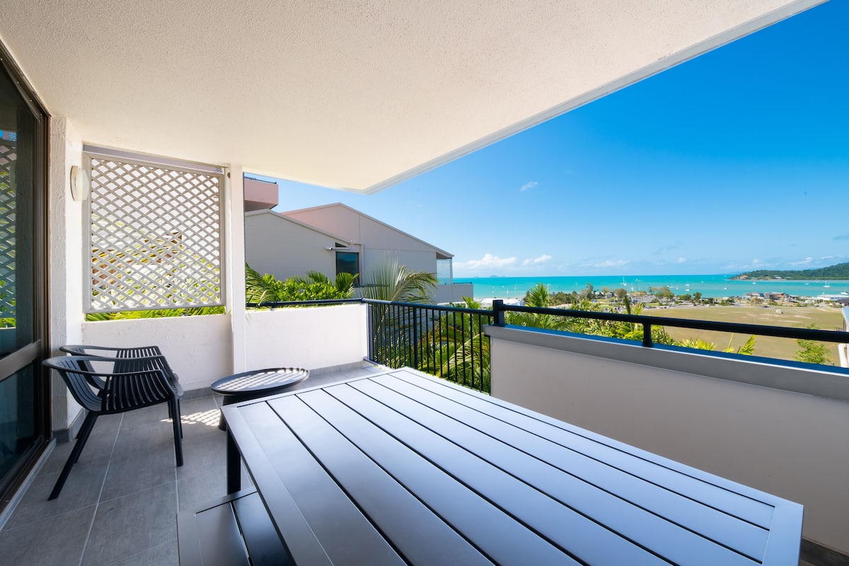 A balcony with a wooden floor, a table, and two chairs. It overlooks a beach with clear blue water and a distant shoreline.