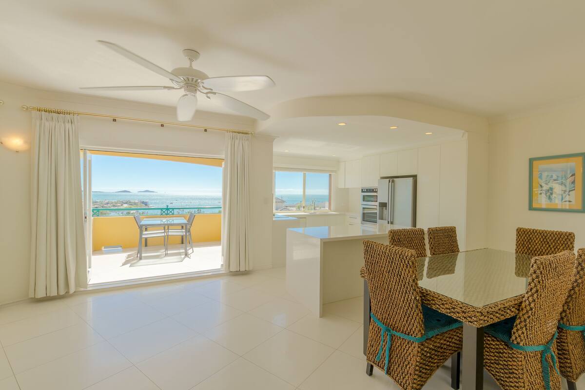 A spacious dining area with a glass table and woven chairs, a ceiling fan, and a view of the ocean through large windows.