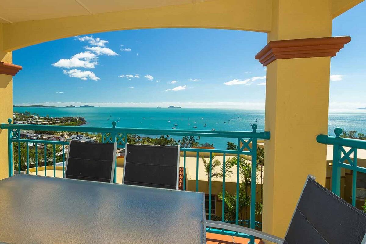 A balcony with a table and chairs, overlooking a coastal view with clear skies and distant islands.