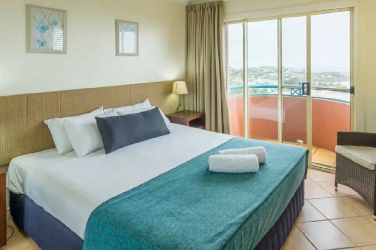A hotel room with a bed, a nightstand, a chair, a balcony with a view, and a window.