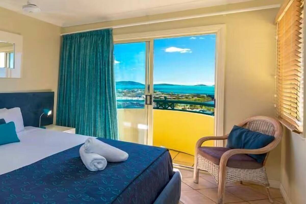 A hotel room with a bed, a chair, a balcony with a view, and a window with blinds.
