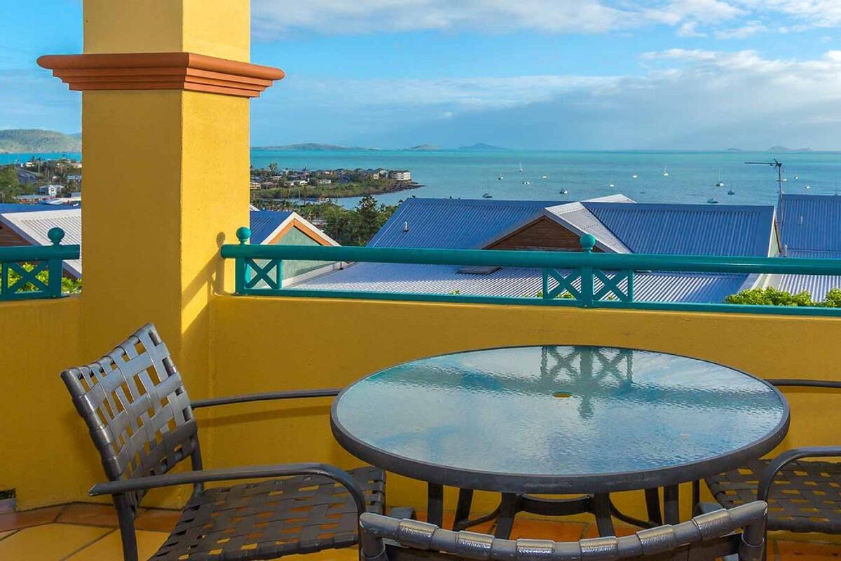 A balcony with a glass table and two chairs, overlooking a coastal view with buildings and boats.