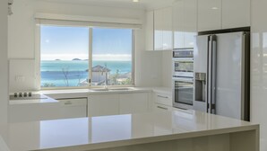 A modern kitchen with white cabinetry, a stainless steel refrigerator, and a large window offering a view of the sea. - Toscana Village Resort (Airlie Beach)