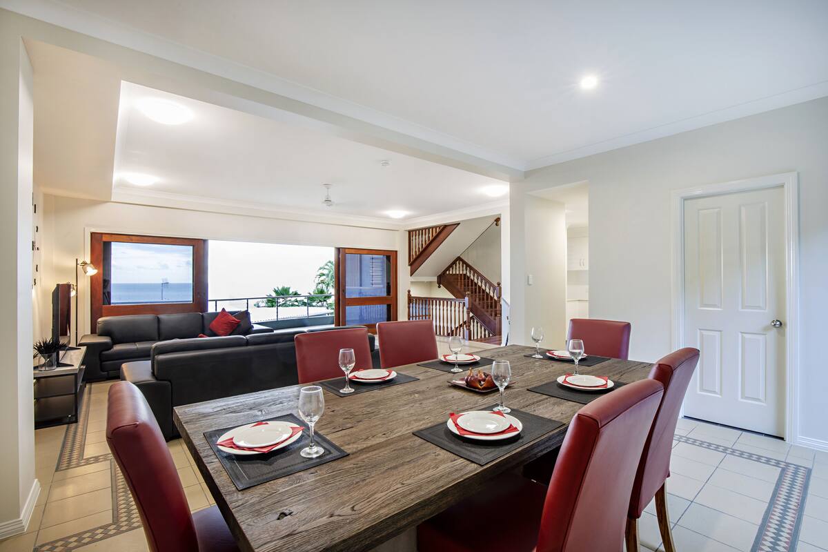 A dining area with a wooden table set for six, surrounded by red chairs, and a living area with a sofa and a view of the outdoors.