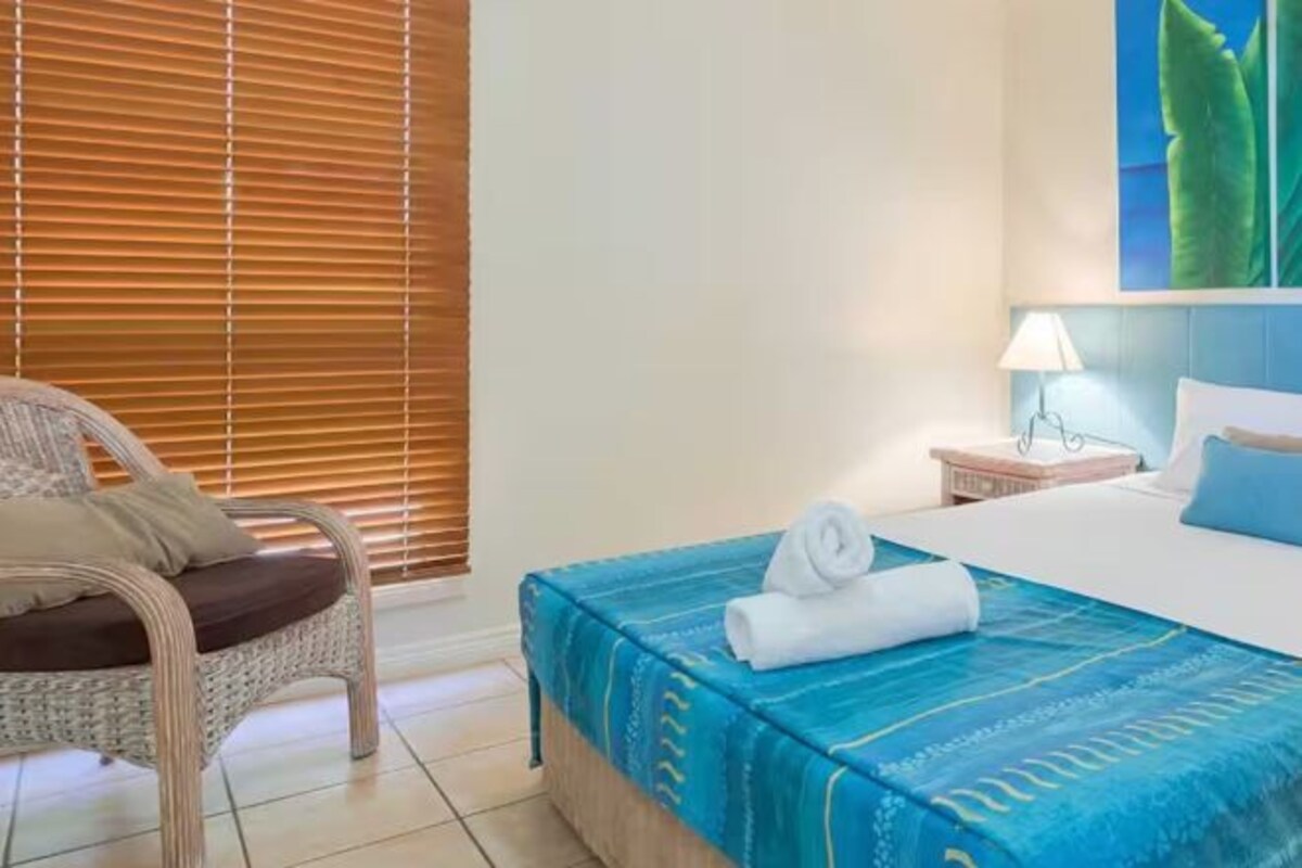 A hotel room with a bed, a chair, a nightstand, and a window with blinds.
