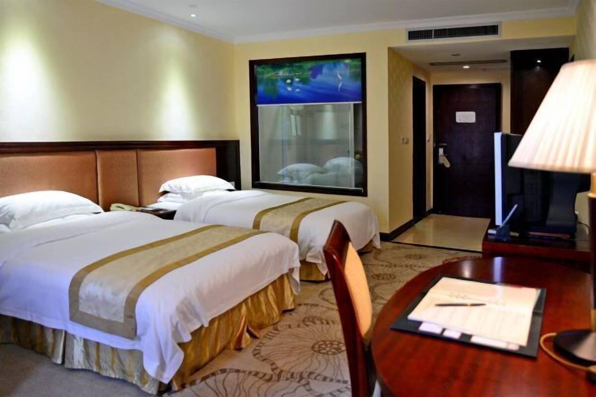A hotel room with two beds, a large window with a view, a desk with a lamp, and a door.