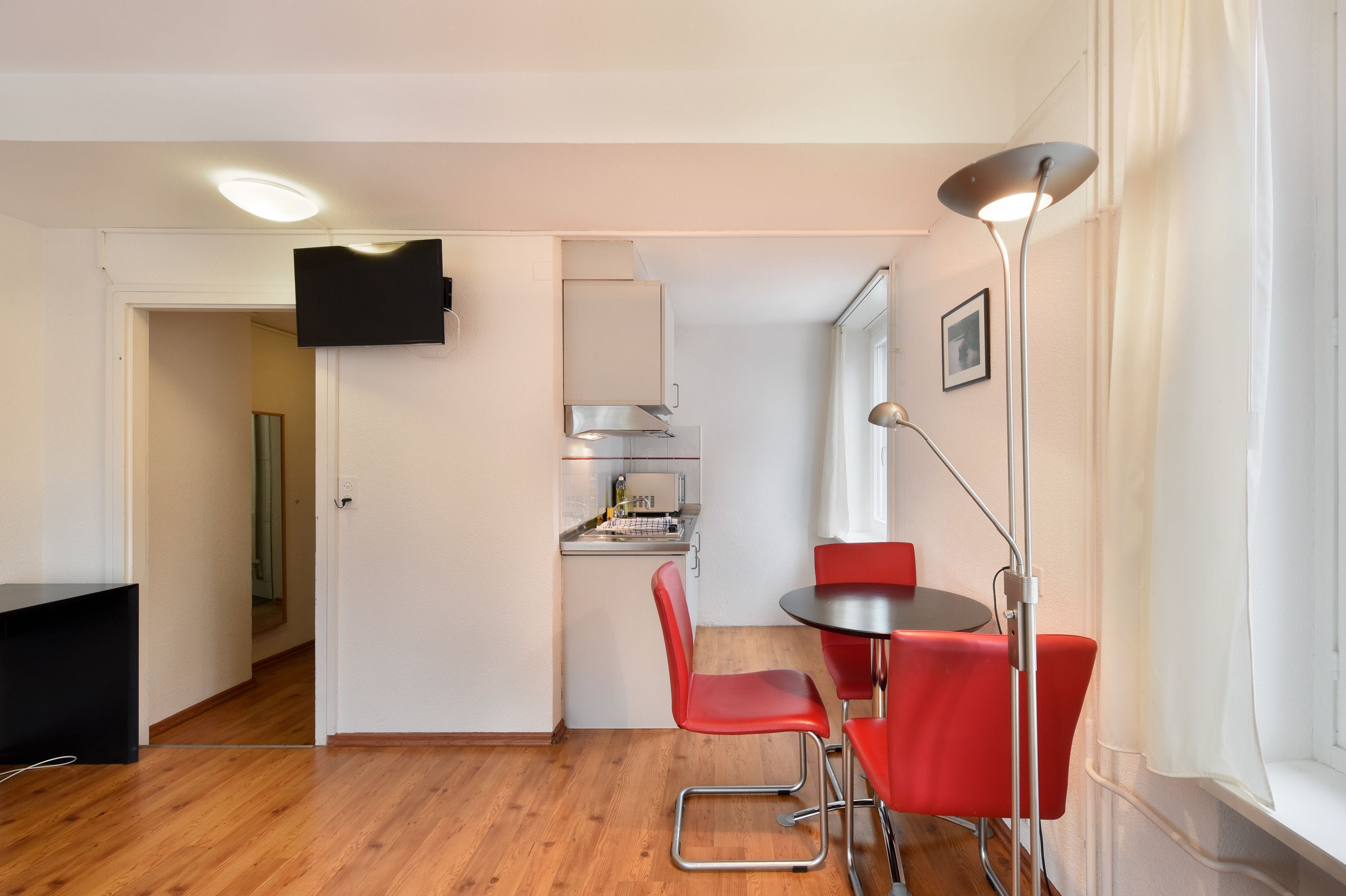 A compact kitchen with modern appliances, a dining area with red chairs, and a flat-screen TV mounted on the wall.